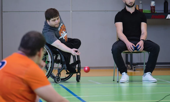 Na zdjęciu dwóch mężczyzn grających w boccia. Jeden z nich rzuca czerwoną kulą. Siedzi na wózku. Drugi - obok siedzi na krześle i trzyma niebieskie kule w rękach. Z boku widzowie.