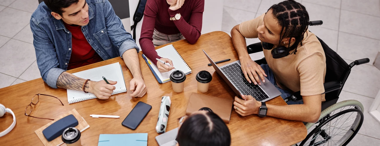 Grupa czterech młodych osób siedzi przy stole i współpracuje nad projektem. Jedna z osób korzysta z laptopa, inna pisze w notesie. Na stole leżą zeszyty, kubki z kawą i smartfony. Jedna z osób porusza się na wózku inwalidzkim.