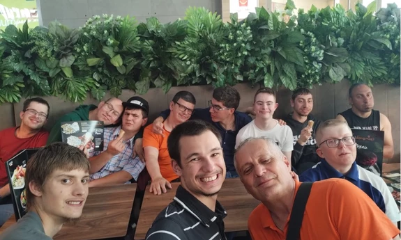 Mężczyźni z niepełnosprawnością wraz z trenerem mgr Januszem Cyranem. Przebywają w restauracji. Na twarzach zadowolenie i uśmiechy.