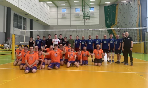 Na zdjęciu pracownicy i studenci UBB na hali sportowej. Stoją na boisku do gry w siatkę. W pierwszym rzędzie klęczą osoby w pomarańczowych koszulkach. Są to osoby z niepełnosprawnością intelektualną z sekcji zunifikowanej siatkówki specjalnej.