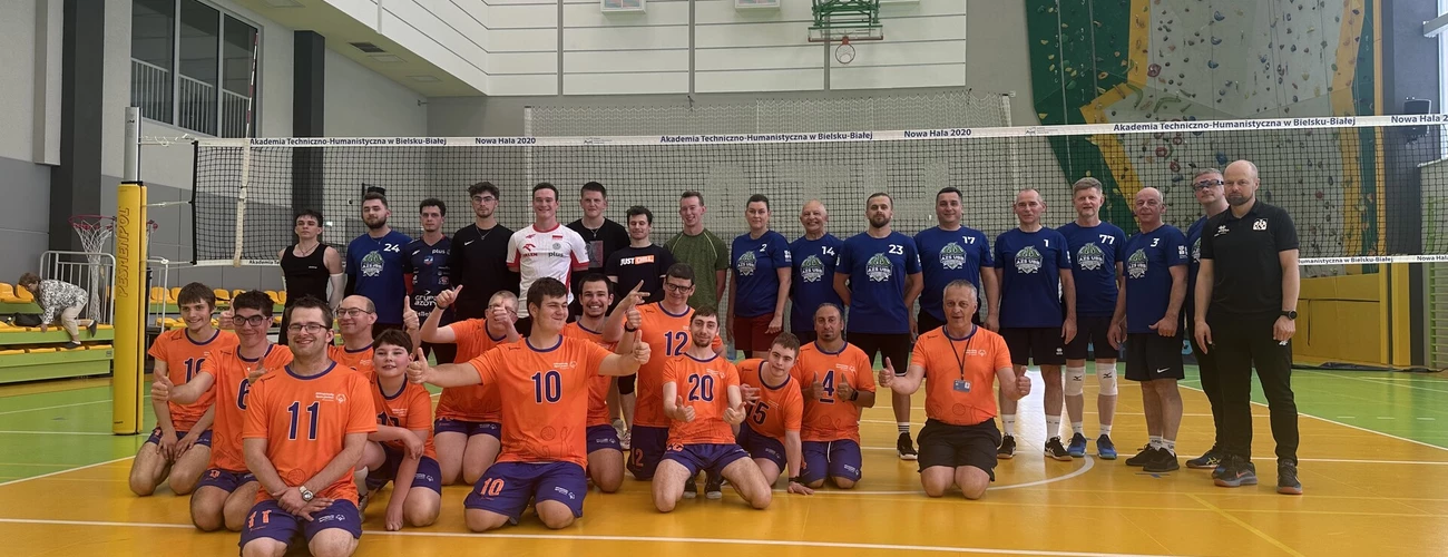 Na zdjęciu pracownicy i studenci UBB na hali sportowej. Stoją na boisku do gry w siatkę. W pierwszym rzędzie klęczą osoby w pomarańczowych koszulkach. Są to osoby z niepełnosprawnością intelektualną z sekcji zunifikowanej siatkówki specjalnej.